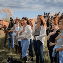 A group of women stand in a grassy field with horses behind them, each with one hand on their heart and one on their abdomen, eyes closed, participating in a peaceful outdoor activity. A dog is in the foreground.